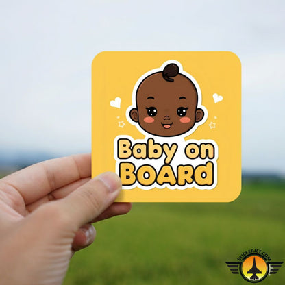 Hand holding a yellow Baby On Board Sticker – Playful Baby Face Design with a cartoon baby face against a blurred natural background.