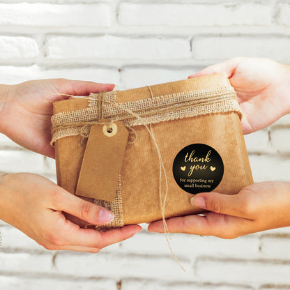 Two hands holding a small brown gift box with a Thank You Sticker – Black & Gold Design tag against a white brick wall.