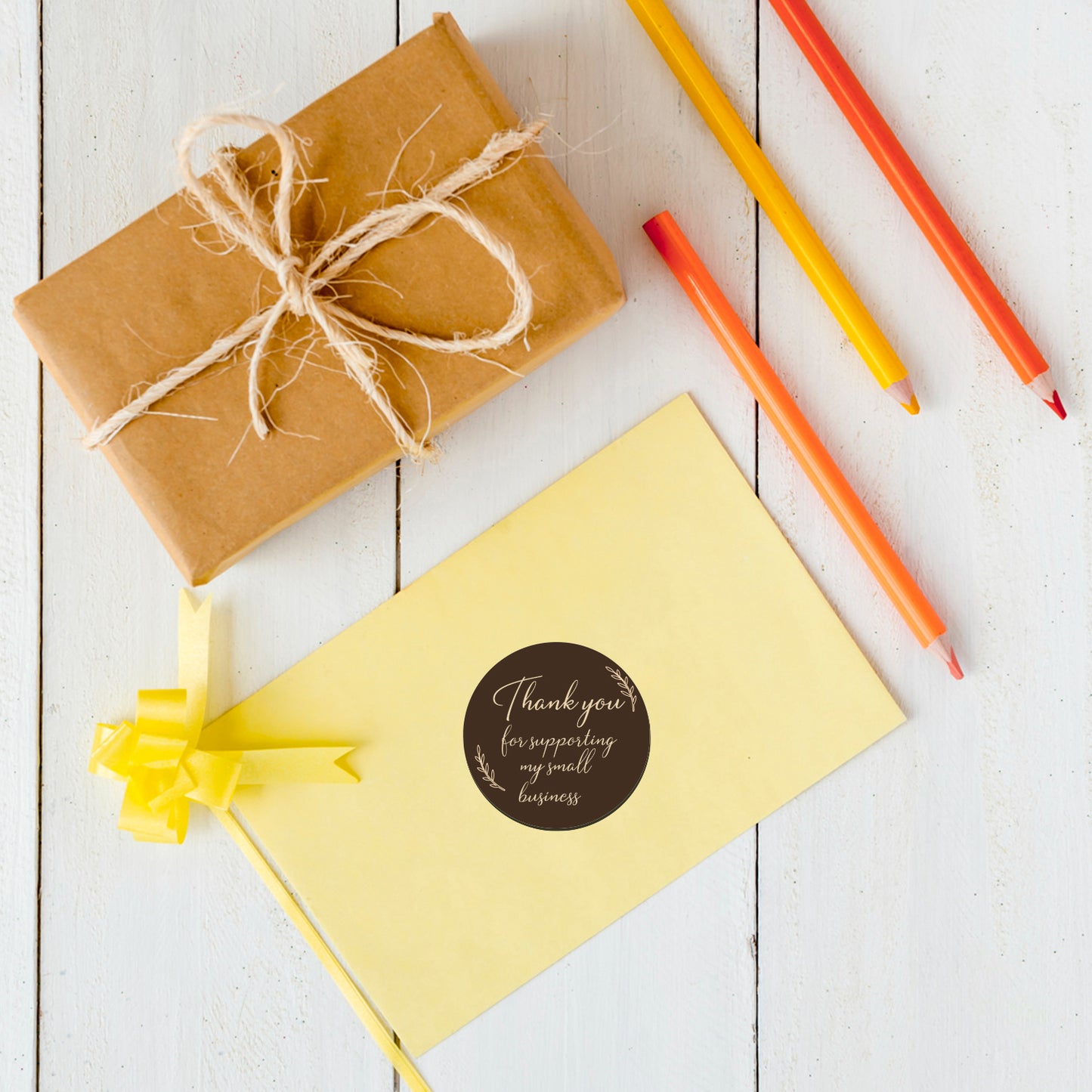 Gift box with brown paper and string, yellow card having Thank You Sticker – Brown Script Design with ribbon, and colored pencils on a white wooden surface.